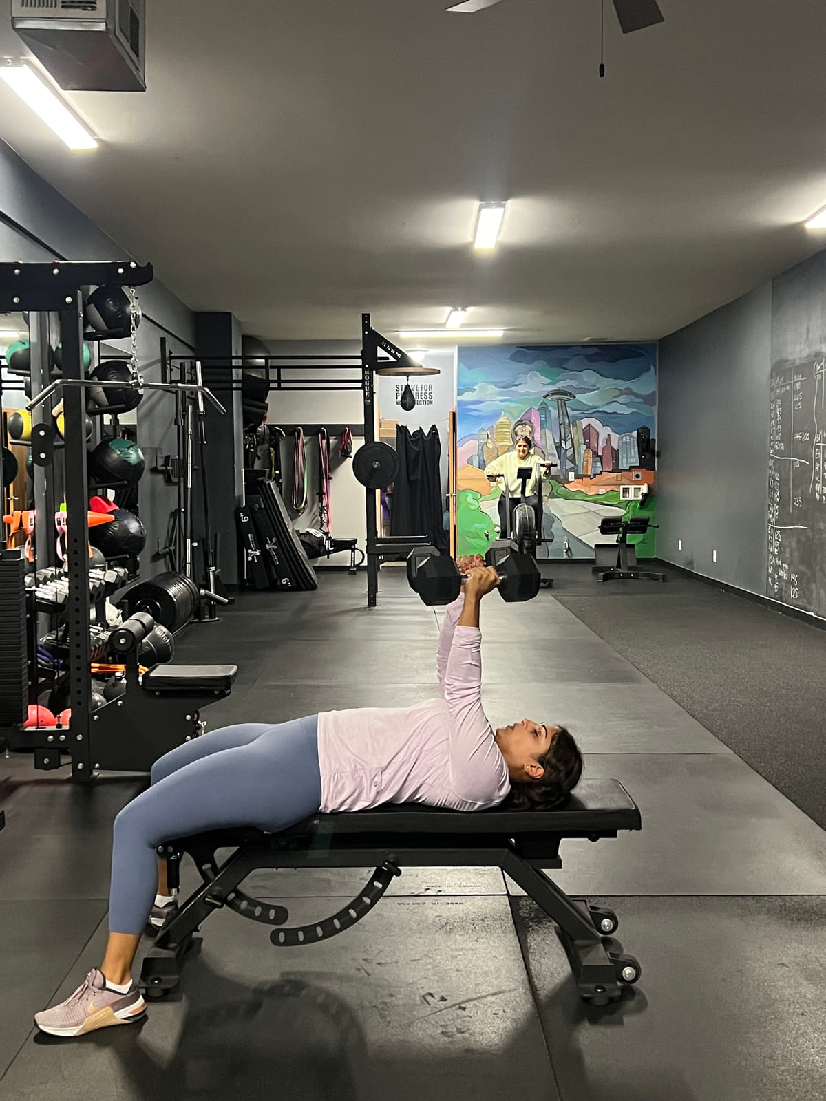 Dumbbell bench press on the main floor at Seattle Muscle, with the Seattle skyline mural on the back wall